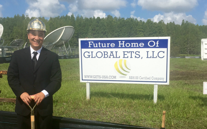 GETS CEO Dan Tang in a suit standing in front of a signboard that reads 'Future Home Of GLOBAL ETS, LLC' with satellite dishes in the background and a forested area behind under a clear sky.