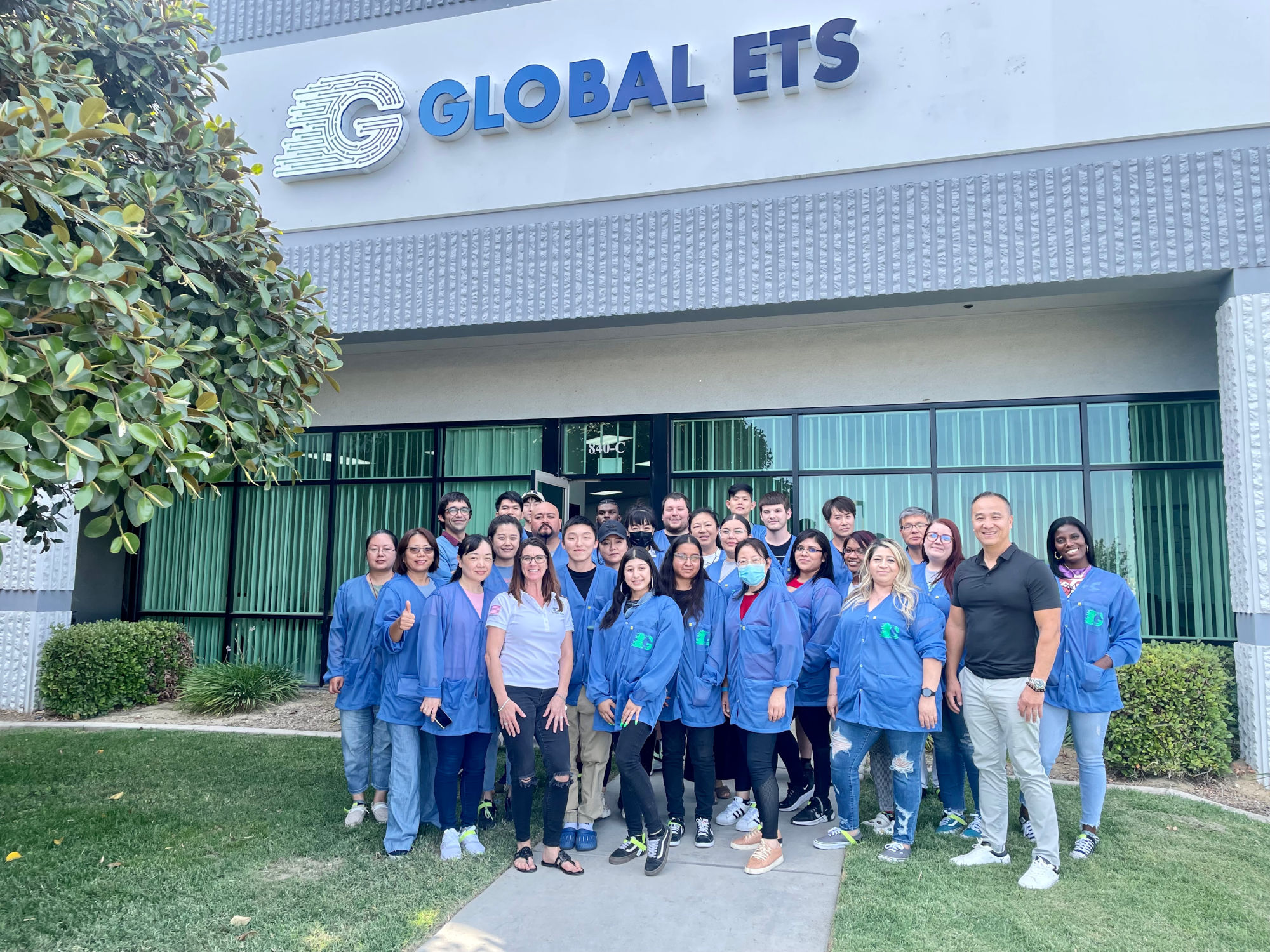 A group of Global ETS employees wearing blue lab coats outside in front of a building with a sign that reads 'GLOBAL ETS.' They are gathered on a lawn next to a bush and in front of windows with vertical blinds.