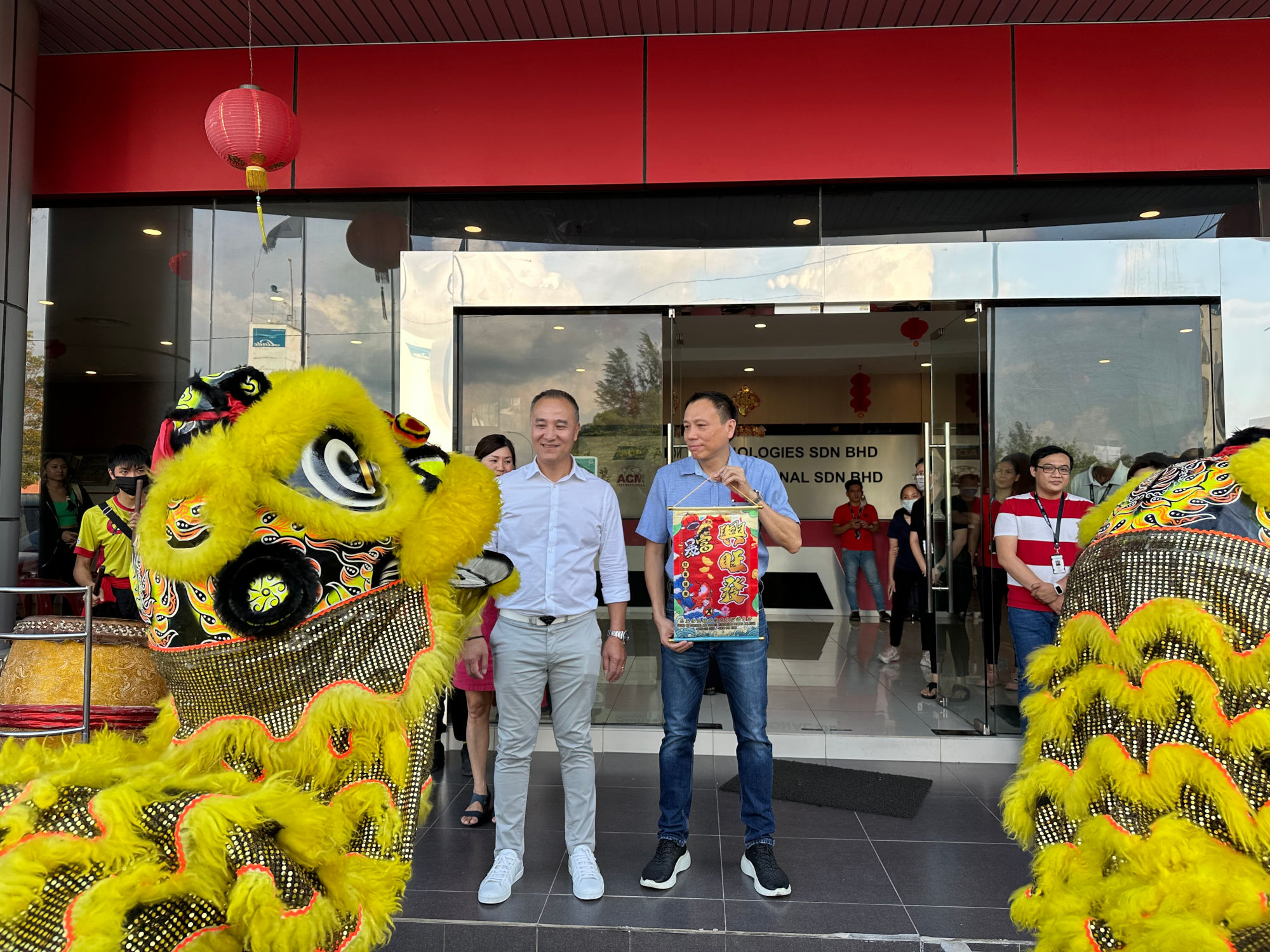 A colorful traditional lion dance performance outside a building with a red overhang and a lantern hanging above. Two people in lion dance costumes with yellow and black fur are visible. GETS CEO Dan Tang and another employee standing next to the entrance holding a red decorative item with Chinese patterns. Onlookers gather at the entrance of the building.