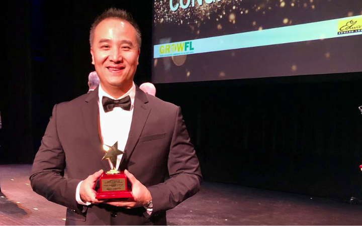 Global ETS, LLC CEO Dan Tang in a black suit and bow tie holding an award for top companies to watch in Florida on a stage with a large screen in the background displaying the text 'CONGRATULATIONS.'