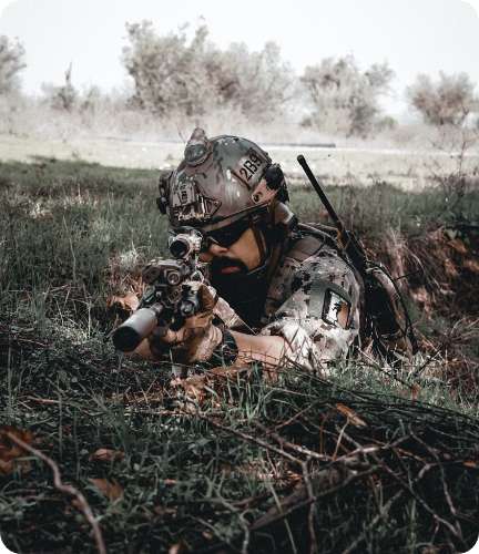 A soldier in camouflage gear lying in the grass aiming a rifle with a scope to represent the ISO9100/AS9100 standard.