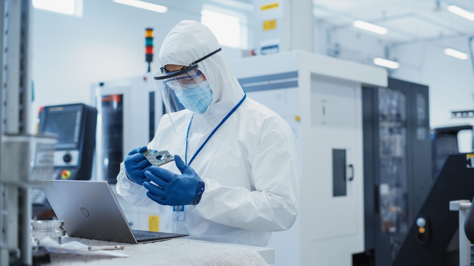 A person in a white protective suit and blue gloves holding an electronic component, standing in a high-tech semiconductor manufacturing facility with machines and equipment in the background.