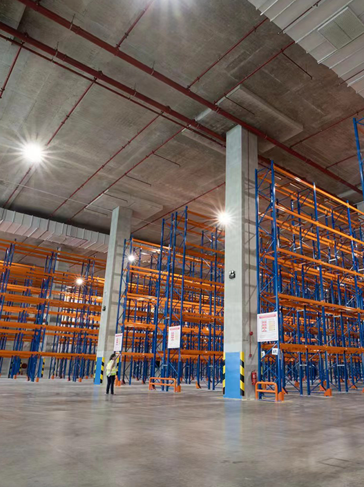 Interior of a large semiconductor warehouse with empty blue and orange industrial shelving units and a high ceiling with bright lights. 