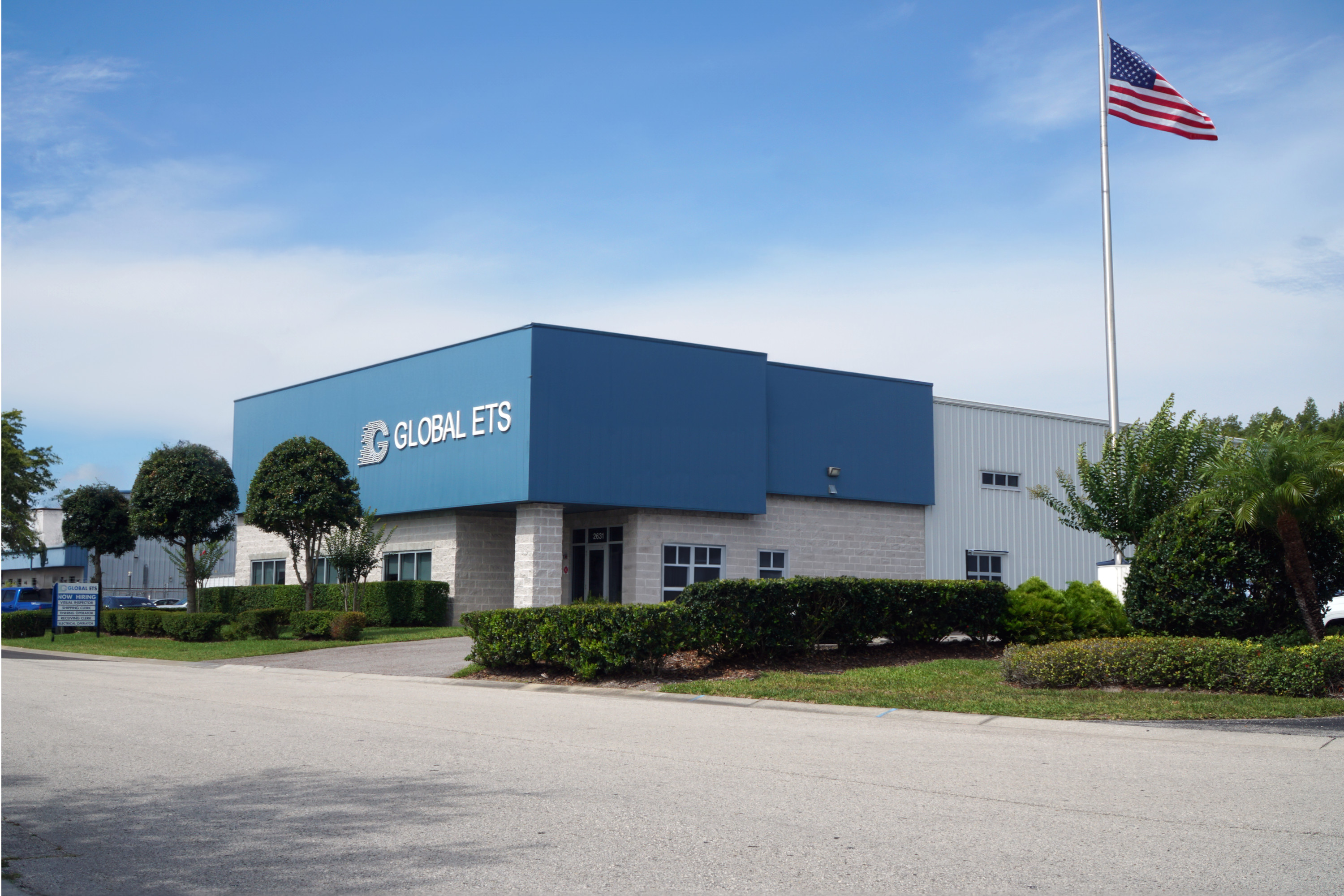 Exterior view of a gray and blue industrial building with a sign reading 'Global ETS' and an American flag flying on a pole. 