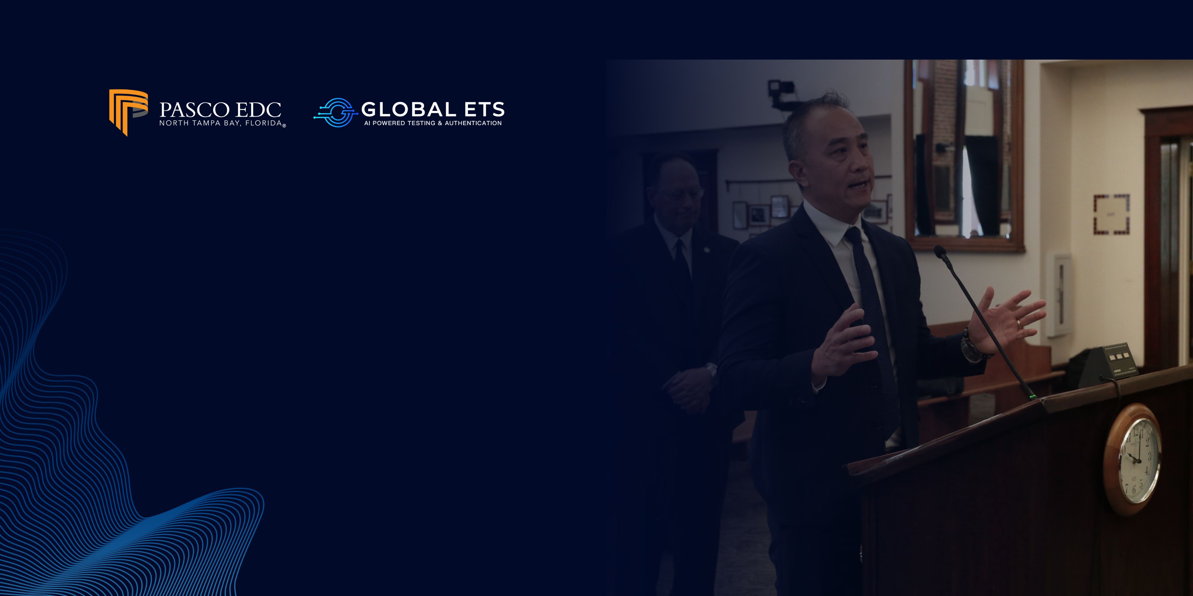 CEO of Global ETS, Dan Tang, in a dark suit is speaking at a podium in a room with wood-paneled walls and a clock, with the logos of Pasco EDC and Global ETS visible on a dark blue background with a wave-like design on the left side of the image.