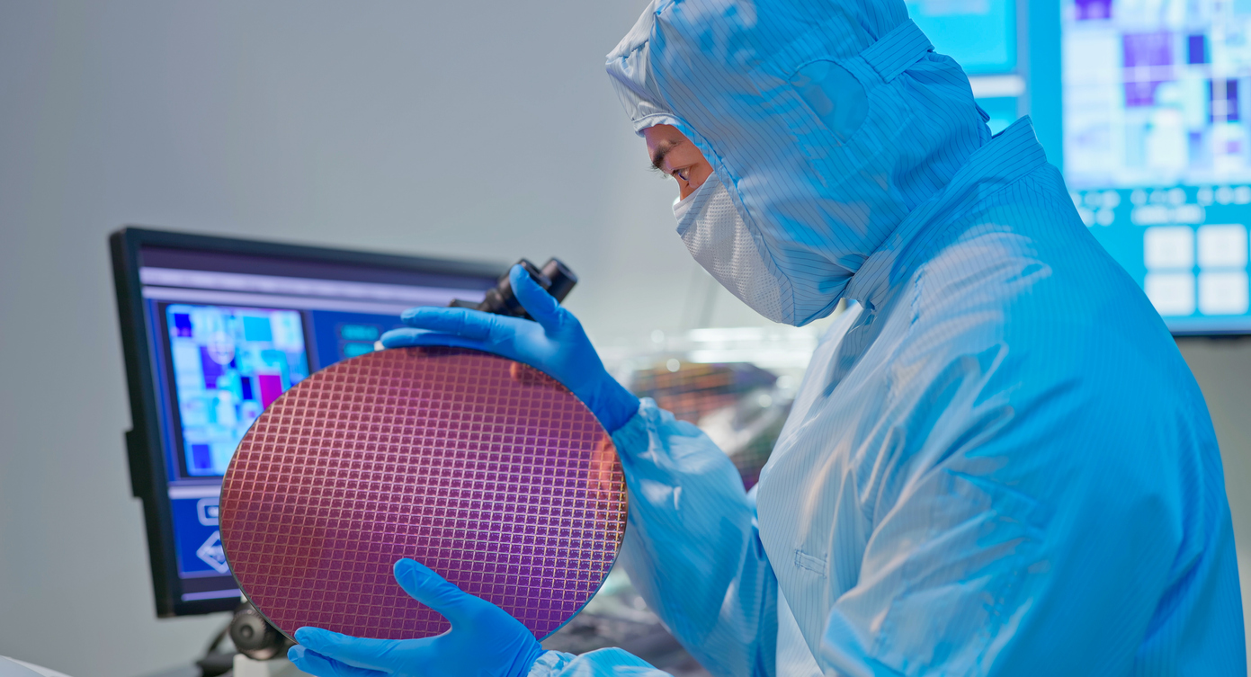 A person in a cleanroom suit holding a silicon wafer in a semiconductor manufacturing facility, with monitoring screens displaying graphs and data in the background. 