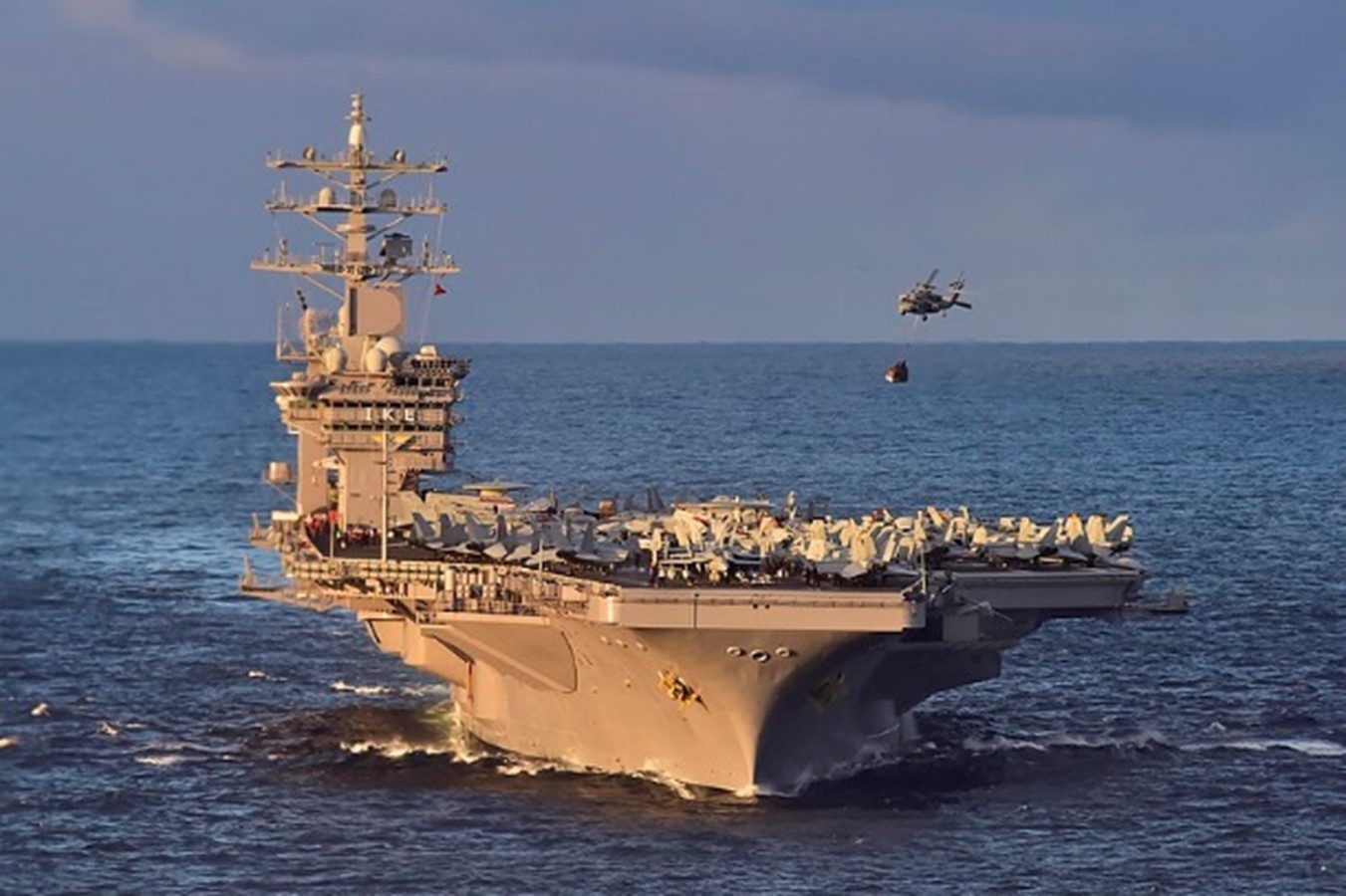 A military aircraft carrier sailing on the ocean with a helicopter flying nearby.