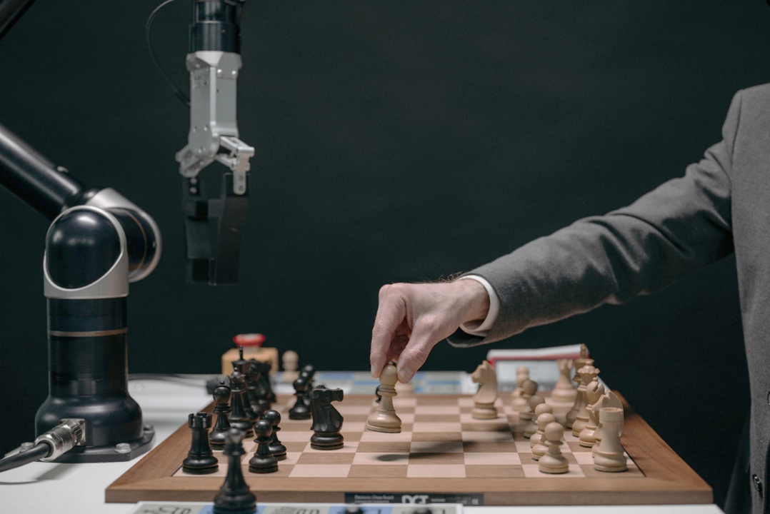 A human hand moving a white chess piece on a chessboard while playing against a robotic arm running on artificial intelligence. 