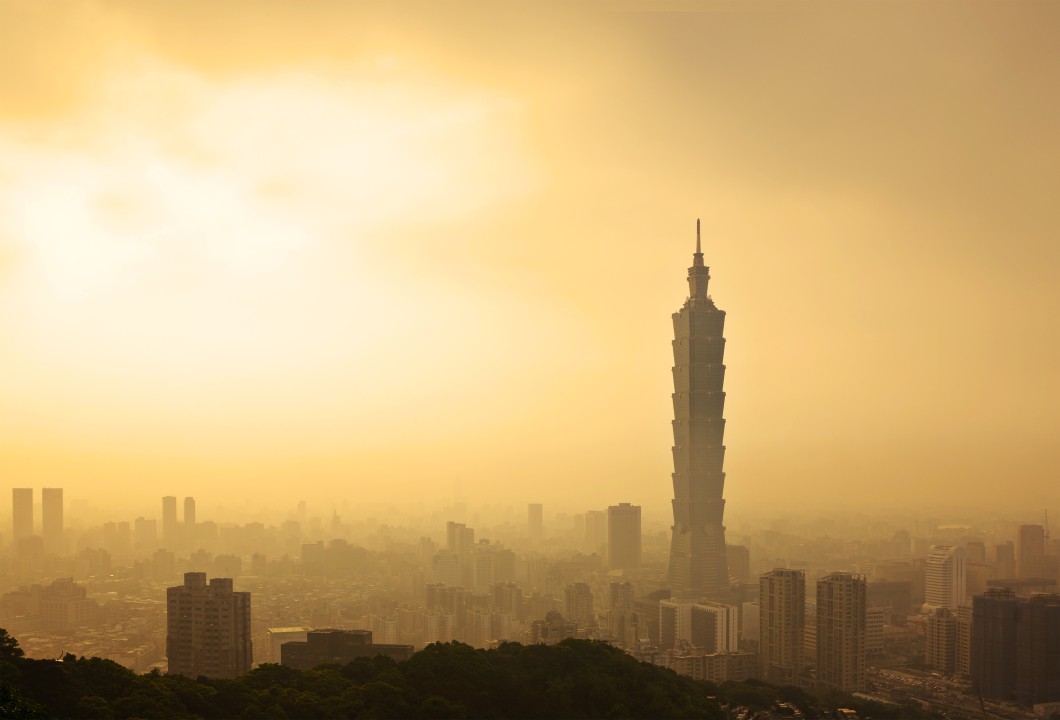 A hazy golden sunset over a cityscape with the silhouette of Taipei 101 skyscraper towering over the surrounding buildings.