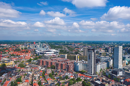 Aerial view of The Netherlands with a mix of modern skyscrapers and traditional red brick buildings, a large stadium in the background, and a blue sky with scattered clouds.