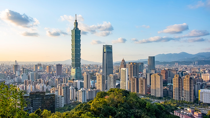 A panoramic view of the Taipei city skyline with the Taipei 101 skyscraper standing tall surrounded by numerous other buildings, set against a backdrop of lush green mountains and a clear blue sky with clouds. 