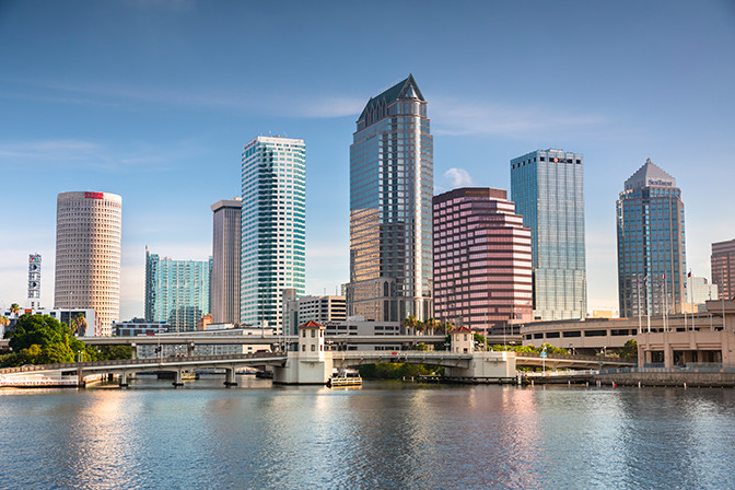 Skyline of Tampa with various high-rise buildings reflected in a calm river under a clear sky.