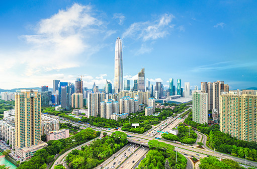 Aerial view of Shenzhen with tall skyscrapers, a prominent central tower, green trees, and a multi-lane highway with cars. 