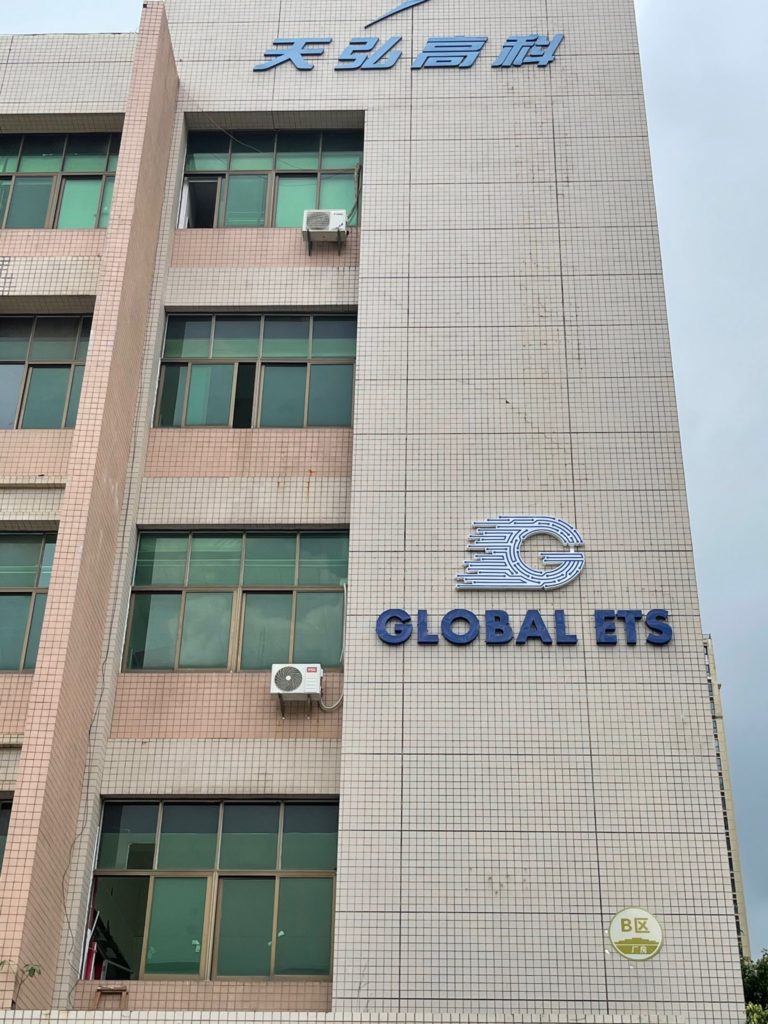 Tall beige building with large windows and two air conditioning units on the exterior. The building has signage that reads 'Global ETS' in blue letters with a circular logo above it. There are also Chinese characters on the top and a green circular logo on the bottom right corner. 