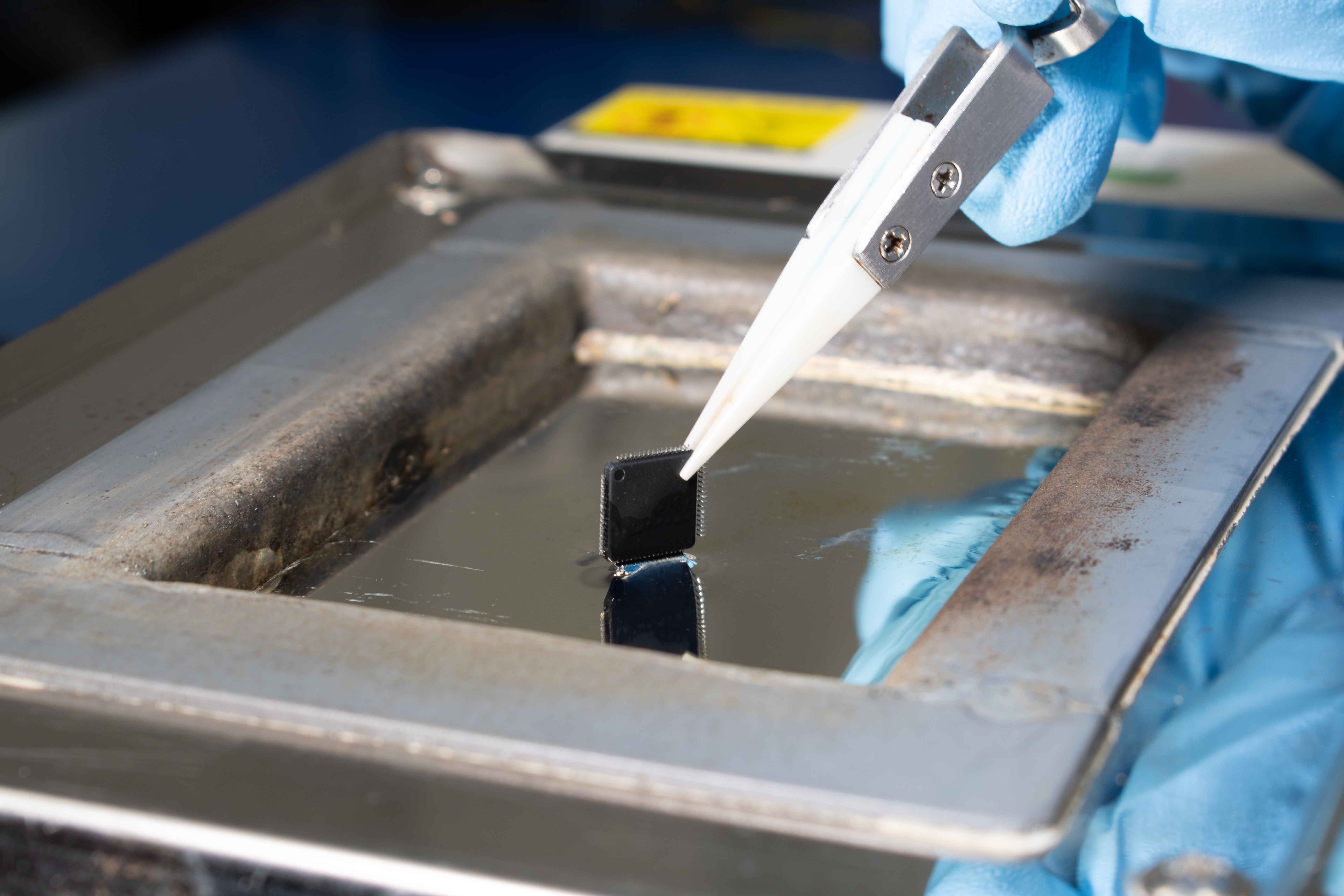 A Global ETS technician wearing blue gloves using tweezers to perform re-tinning on an electronic component using a solder pot.