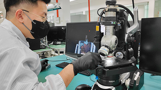 A Global ETS technician in a laboratory coat operating a microscope to view the die of semiconductors, surrounded by various pieces of component testing equipment and monitors displaying magnified images.