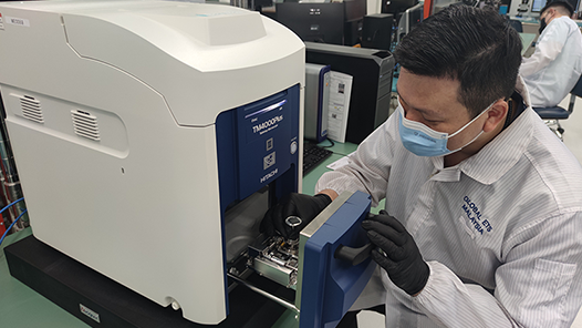 A Global ETS technician wearing gloves and a lab coat with the words “Global ETS” stitched near the collar, is working with a blue and white semiconductor testing machine in a laboratory setting, with computers and equipment in the background.